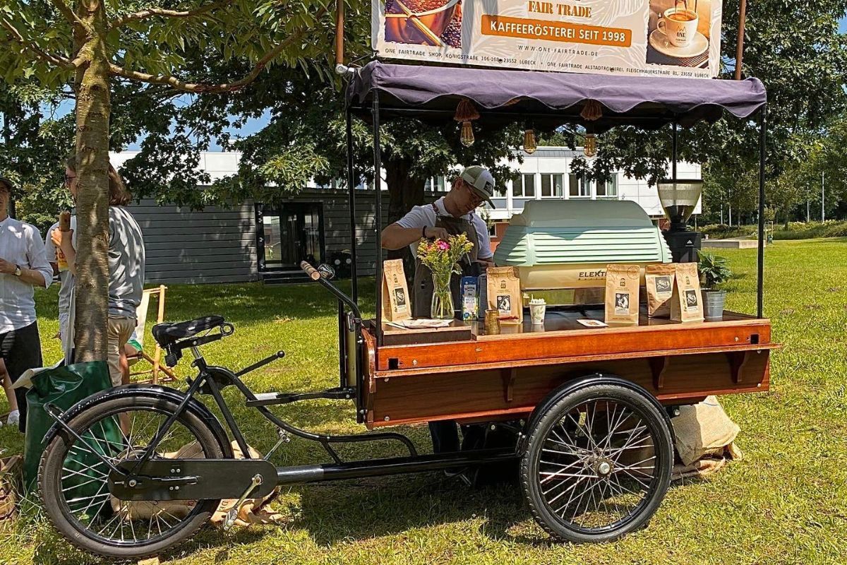Coffee-Bike von ONE Fairtrade auf Rasen bei Outdoor-Event, Mitarbeiter bereitet Fairtrade Bio Kaffeespezialitäten als Barista zu